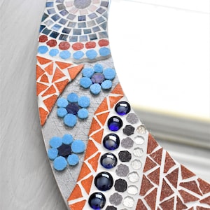 May include: A round mirror with a mosaic frame. The frame is made of small, colorful tiles in shades of blue, red, orange, and gray. The mirror is surrounded by a border of blue flowers.