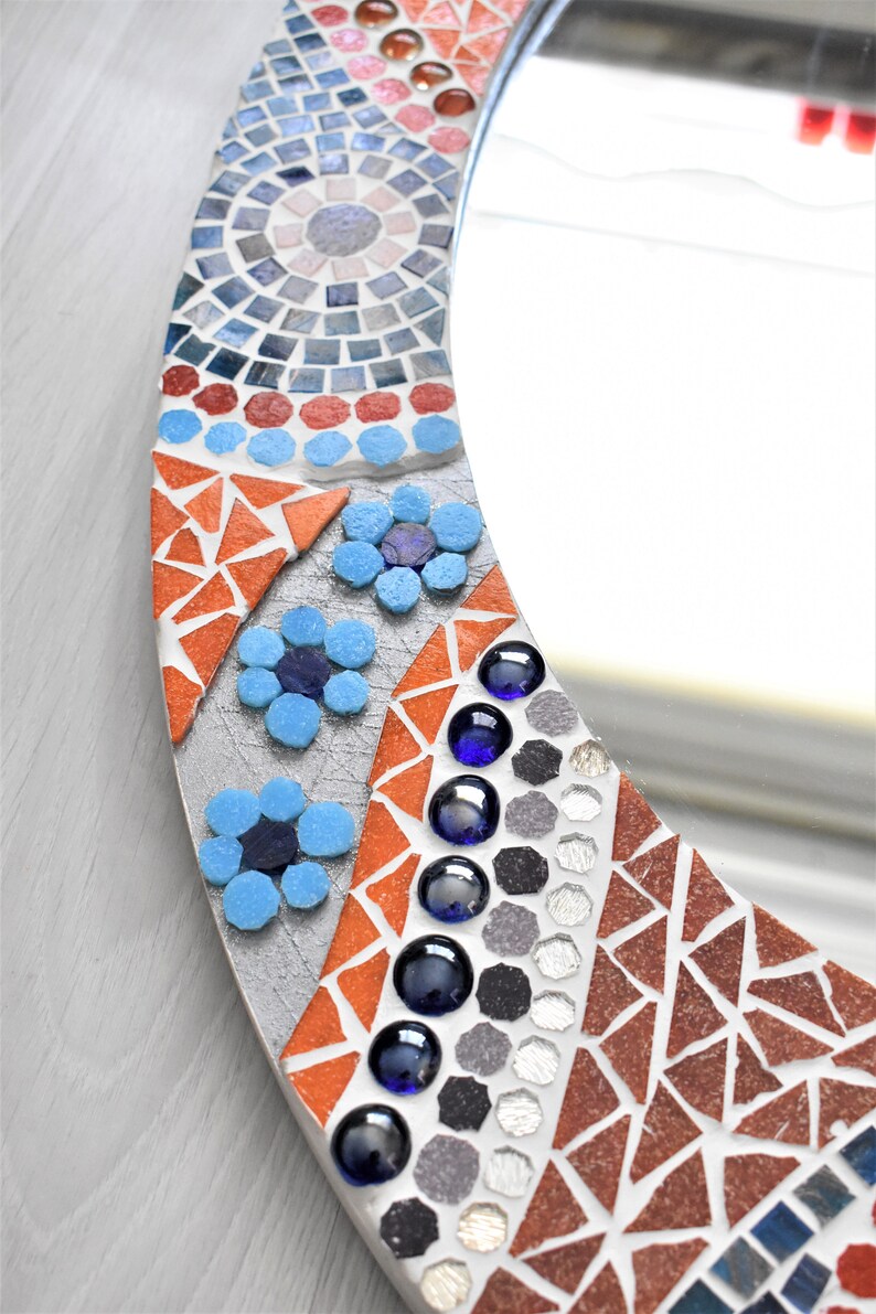 May include: A round mirror with a mosaic frame. The frame is made of small, colorful tiles in shades of blue, red, orange, and gray. The mirror is surrounded by a border of blue flowers.