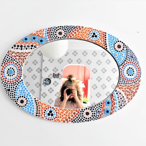 May include: Oval mirror with a mosaic frame in shades of blue, orange, and brown. The frame features geometric patterns and circles.