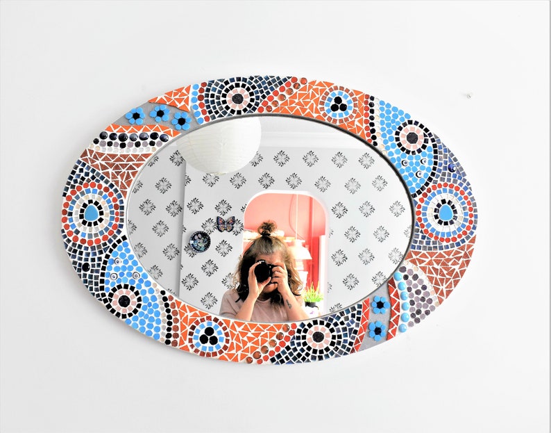 May include: Oval mirror with a mosaic frame in shades of blue, orange, and brown. The frame features geometric patterns and circles.