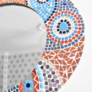 May include: A round mirror with a mosaic design in shades of blue, red, orange, and white. The mosaic features geometric shapes and patterns, including circles, triangles, and squares.