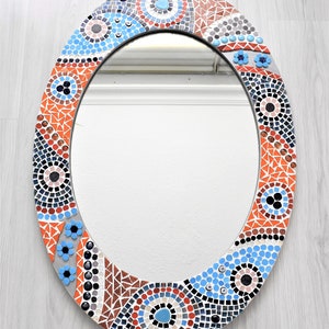 May include: Oval mirror with a mosaic frame. The frame is made of small, colorful tiles in shades of blue, red, orange, and black. The tiles are arranged in a geometric pattern.