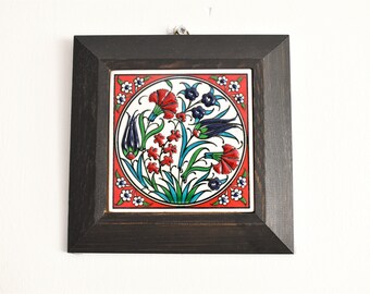 May include: A framed ceramic tile with a colorful floral design. The tile features a white background with red, blue, and green flowers and leaves. The frame is black wood.
