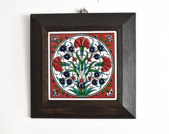 May include: A framed ceramic tile with a red, white, blue, and green floral design. The tile features a circular pattern of stylized carnations and leaves.