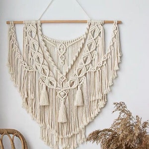 Op de afbeelding: Een grote, off-white macramee wandhanger met een geometrisch patroon en franjes. De hanger is gemaakt van katoenen koord en heeft een houten lat aan de bovenkant.