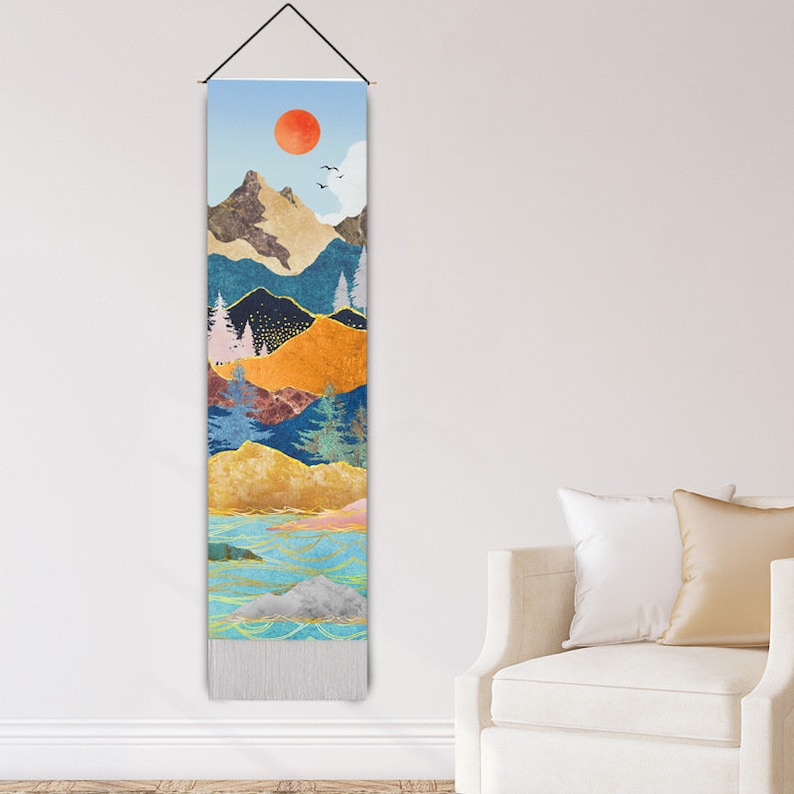 Puede incluir: Un tapiz de pared vertical con un dise&ntilde;o de paisaje. La obra de arte presenta un sol rojo, monta&ntilde;as, &aacute;rboles y una masa de agua. La paleta de colores incluye tonos azules, dorados y marrones. La parte inferior del tapiz tiene flecos.