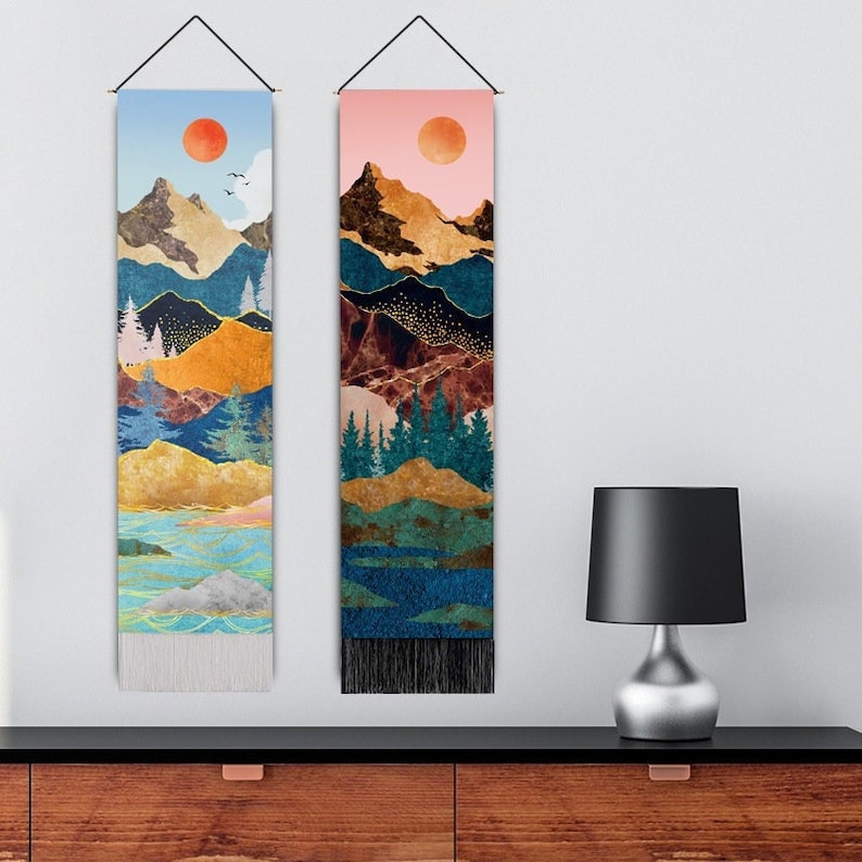 Puede incluir: Dos tapices de pared verticales con dise&ntilde;os de paisajes de monta&ntilde;a. Uno presenta una combinaci&oacute;n de colores azul y naranja con un sol rojo, el otro una combinaci&oacute;n de colores rosa y marr&oacute;n con un sol rojo. Ambos tienen un cord&oacute;n negro para colgar y flecos.