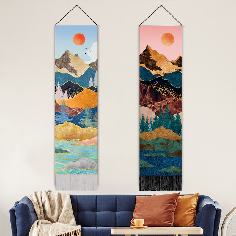 Puede incluir: Dos tapices verticales con dise&ntilde;os de paisajes de monta&ntilde;a. Uno presenta un cielo azul con un sol rojo, el otro un cielo rosa. Ambos tienen cadenas monta&ntilde;osas doradas, azules y marrones, &aacute;rboles y una franja en la parte inferior. Colgados de varillas met&aacute;licas negras.