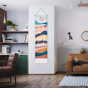 May include: A tapestry wall hanging with a colorful abstract landscape design. The tapestry features a layered mountain scene with a sun and trees in the center. The tapestry has a fringe border.