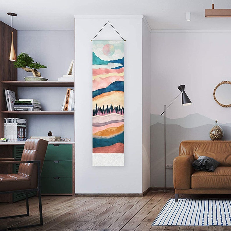 May include: A tapestry wall hanging with a colorful abstract landscape design. The tapestry features a layered mountain scene with a sun and trees in the center. The tapestry has a fringe border.