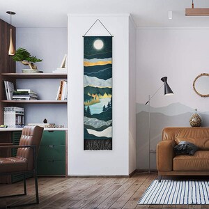 May include: A tapestry wall hanging with a dark blue, green, and gold mountain landscape design. The tapestry features a full moon and a lake with trees. The tapestry has a fringe border.