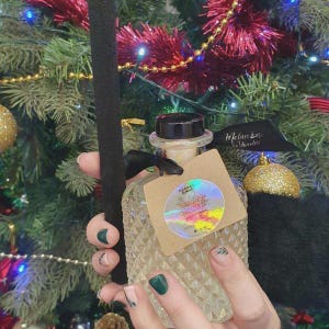 May include: A clear glass bottle with a diamond pattern, black cap, and a small tag. The bottle contains a light-coloured liquid and is held in front of a Christmas tree. A black ribbon is tied around the neck of the bottle.