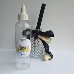 May include: A clear plastic squeeze bottle with a white nozzle cap and a label reading Flying Fish, Eucalyptus, Lemon and Wild Mint, alongside a glass diffuser bottle with a textured surface, a black ribbon, a black reed, and a cork stopper.