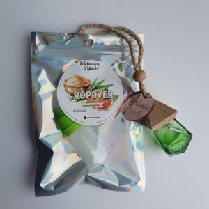 Può includere: Una bustina olografica contenente un piccolo flacone bianco con tappo verde e un'etichetta rotonda con il testo CROPOVER, pompelmo e canna da zucchero. È attaccato un piccolo flacone in vetro verde con tappo in legno e targhetta.