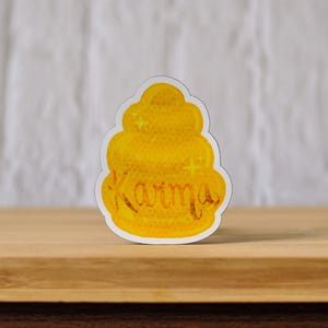 May include: A yellow and white sticker with the word "Karma" written on it. The sticker is shaped like a jar of honey.