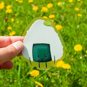 Könnte beinhalten: Ein weißer und grüner Cartoon-Onigiri-Aufkleber mit einem lächelnden Gesicht. Der Aufkleber befindet sich auf einem grünen Hintergrund mit gelben Blumen.