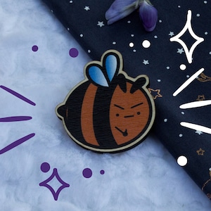 Peut inclure: Une épingle en bois en forme d'abeille avec un corps rayé noir et brun, des ailes bleues et une expression boudeuse. L'épingle est sur un fond blanc avec des accents violets et un fond noir avec des étoiles blanches et un cerf doré.