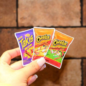 May include: Three snack chip bags, two are Cheetos, one is Takis. The Cheetos bags are orange and red with the text "Cheetos Flamin' Hot" and "Cheetos Flamin' Hot Limon". The Takis bag is purple and red with the text "Takis Fuego".