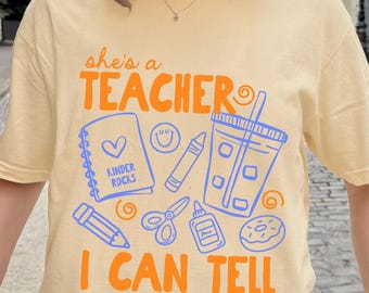 May include: A pale yellow short-sleeve t-shirt featuring orange text that reads "she's a TEACHER I CAN TELL" and blue line art graphics of a spiral notebook labeled "KINDER ROCKS", a smiley face, a crayon, a drink cup, a pencil, scissors, glue, and a donut.