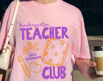 May include: A light pink t-shirt with the words "kindergarten TEACHER ABC CHAOS COORDINATOR CLUB" in purple and orange. The design includes a pencil, apple, and notebook. The shirt is short-sleeved.