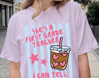 May include: A light purple t-shirt featuring a graphic with vertical light blue stripes, two pink stars, and a brown iced coffee cup with pink hearts. The text reads, "She's A FIRST GRADE TEACHER I CAN TELL" in pink lettering.