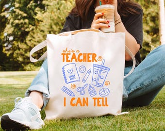May include: A cream-colored canvas tote bag featuring orange text that reads "she's a teacher I can tell" and blue line art illustrations of a spiral notebook labeled "KINDER ROCKS," a crayon, a tumbler, a pencil, scissors, glue, and a donut.
