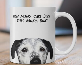 May include: White ceramic mug with a black handle featuring a dog's face and the text "How Many Cups Does This Make, Dad?" The dog has black ears and brown eyes. The mug is on a wooden surface.