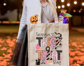 May include: A beige canvas tote bag featuring the text Georgia's TRICK OR TREAT Bag in black, pink, and orange lettering. The design includes illustrations of a black cat, a spider web, a witch hat, a pink pumpkin, and various candy pieces.