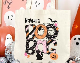 May include: A cream-colored canvas tote bag featuring the text Randi's BOO BAG in large, decorative letters. The design includes black, orange, and pink patterns, a spider, a spiderweb, a black cat, bats, stars, and a small orange jack-o'-lantern.