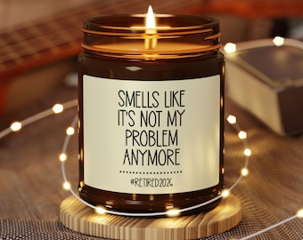 May include: A lit brown glass jar candle with a white label that reads "Smells Like It's Not My Problem Anymore" and "#Retired2026". The candle sits on a wooden coaster, surrounded by string lights.