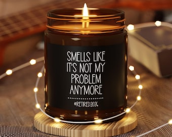 May include: A lit candle in a brown glass jar with a black label. The label reads "Smells Like It's Not My Problem Anymore" and "#Retired2026" in white text. The candle sits on a wooden coaster, surrounded by string lights.