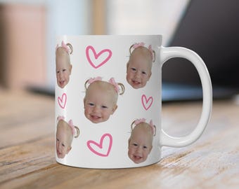 May include: A white ceramic mug featuring a repeating pattern of a smiling toddler's face with blonde hair in pigtails and pink bows, interspersed with hand-drawn style pink heart outlines printed across the exterior surface of the mug.