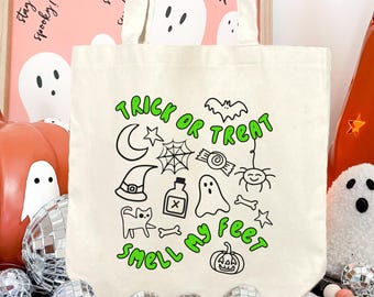May include: A cream-colored canvas tote bag featuring a green and black graphic print with the text "TRICK OR TREAT SMELL MY FEET" surrounded by illustrations of a crescent moon, spider web, bat, candy, witch hat, potion bottle, ghost, cat, and pumpkin.