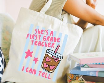 May include: A cream-colored canvas tote bag featuring a graphic print with light blue vertical stripes. The text reads "SHE'S A FIRST GRADE TEACHER I CAN TELL" in pink letters, accompanied by two pink stars and an iced coffee cup with heart shapes.