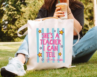May include: A white canvas tote bag featuring vertical light blue stripes and the pink text "SHE'S A TEACHER I CAN TELL" surrounded by yellow and blue star graphics. The bottom right corner includes the cursive signature "Mrs. Griffin".