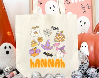 May include: A cream-colored canvas tote bag featuring a colorful Halloween-themed graphic print. The design includes ghosts, a spider web, a pumpkin, a witch hat, and candy, with the name "hannah" printed in large orange letters at the bottom.