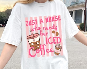 May include: White t-shirt with a pink and white striped design. The shirt features the text "JUST A NURSE who needs her ICED Coffee" in pink, along with illustrations of iced coffee cups and coffee beans. A casual and comfortable tee.