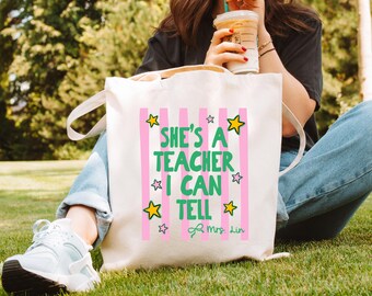 May include: A cream-colored canvas tote bag featuring vertical light pink stripes and the green text "SHE'S A TEACHER I CAN TELL" surrounded by yellow and purple star graphics, with the name "Mrs. Lin" printed in green script at the bottom.