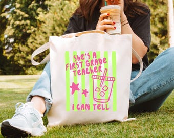 May include: A cream-colored canvas tote bag featuring a graphic design with vertical lime green stripes. The text reads "She's a First Grade Teacher I Can Tell" in pink, accompanied by two pink stars and a pink outline of an iced coffee cup.