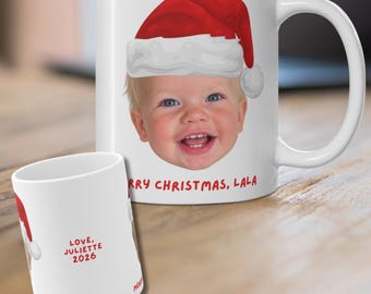 May include: A white ceramic mug featuring a graphic of a smiling child wearing a red Santa hat. The text reads MERRY CHRISTMAS, LALA in red. A smaller view shows the back with the text LOVE, JULIETTE 2026 printed in red on the white surface.