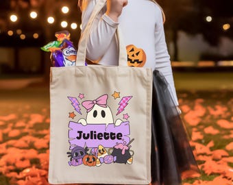 May include: A beige canvas tote bag featuring a graphic of a white ghost wearing a pink bow, holding a purple sign with the name Juliette printed in black. The design includes colorful Halloween elements like a spider, pumpkin, and cauldron.