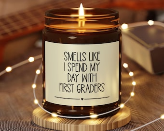 May include: A lit candle in a brown glass jar with a white label. The label reads "Smells Like I Spend My Day With First Graders." The candle sits on a wooden coaster, surrounded by string lights.