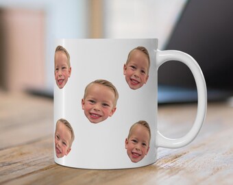 May include: A white ceramic mug featuring a repeating pattern of a young child's smiling face printed across the exterior surface. The mug has a standard cylindrical shape with a curved handle on the right side and a smooth, glossy finish.