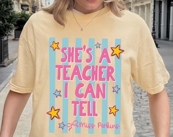 May include: A pale yellow t-shirt featuring a graphic with vertical light blue stripes and the pink text "SHE'S A TEACHER I CAN TELL" followed by "Miss Perkins" in cursive. The design is decorated with yellow and light blue star shapes.