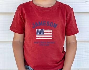 May include: A red short-sleeved t-shirt featuring the name JAMESON in blue text above a graphic of the American flag. Below the flag, the text reads HAPPY 250TH AMERICA 1776-2026 in blue. The shirt is paired with blue denim jeans.
