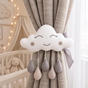 May include: A white plush cloud curtain tieback featuring a smiling face with closed eyes and two light brown circular cheeks. Five felt teardrop-shaped raindrops in shades of grey and beige hang from the cloud by thin white strings.