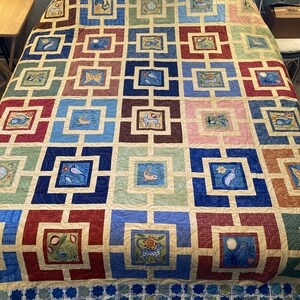 May include: A quilt with a geometric pattern of squares and rectangles in shades of blue, green, red, and brown. Each square features a different design, including birds, butterflies, and flowers. The quilt is edged with a blue and white border.