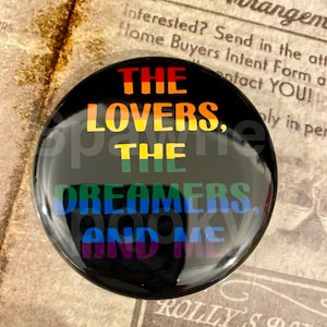 May include: A black round button with the text "THE LOVERS, THE DREAMERS, AND ME" in rainbow colors. The button is glossy and reflects the surrounding environment.