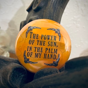 May include: A round, orange button with the text "THE POWER OF THE SUN, IN THE PALM OF MY HAND." The button has a glossy finish and a design of four mechanical wings around the text. The button is a pinback style.