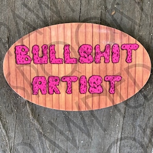 Puede incluir: Cartel de madera ovalado con las palabras "BULLSHIT ARTIST" en letras rosas goteantes. El cartel tiene un fondo de vetas de madera y está sobre una superficie de madera desgastada.